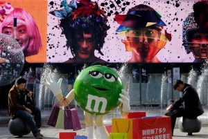 Chinese men browse their mobile phones near a M&M chocolates figure on display at a shopping mall in Beijing, Wednesday, Oct. 28, 2015. Facing pressure to pep up a cooling economy, Chinese leaders met on Monday and will announce a five-year plan effort to shift China from reliance on trade and investment to more self-sustaining growth driven by domestic consumption. The Chinese words on the bag reads "Don't worry how much you shop, just spend to release stress"  (AP Photo/Andy Wong)