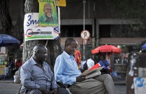 Tanzanians sit next to a tree, underneath an election poster for ruling party presidential candidate John Magufuli, as they await election results in Dar es Salaam, Tanzania Tuesday, Oct. 27, 2015. The presidential race is the most tightly contested since Tanzania became independent in 1961, with the ruling party being challenged by a former member who was recently the country's prime minister. (AP Photo/Khalfan Said)
