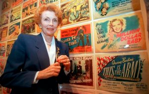 FILE - In this May 14, 1998 file photo, Joan Leslie stands near the movie poster for Irving Berlin's "This is the Army" in which Leslie starred with Ronald Reagan, at the museum on the Warner Bros. lot during a tour marking the film studio's 75th anniversary in Burbank, Calif. Leslie, whose expressive almond eyes and innocent beauty made her one of the most popular film ingenues of the 1930s and 40s, has died at the age of 90. She died in Los Angeles on Monday, Oct. 12, 2015, her family announced in an obituary. (AP Photo/Susan Sterner, File)